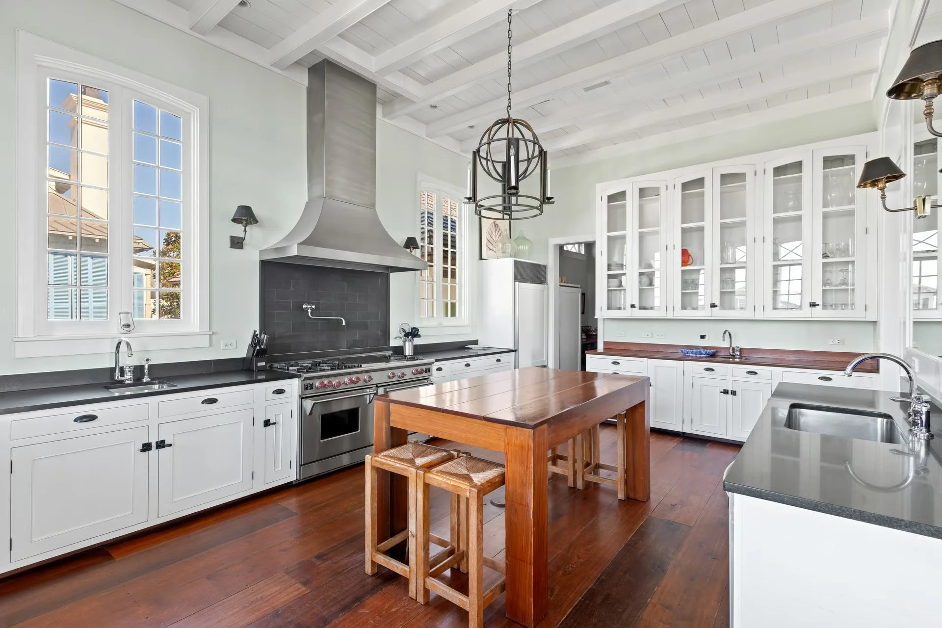 Modern kitchen with white cabinets, black granite countertops, a stainless steel oven and range, a wooden dining table with matching stools, and large windows letting in natural light.