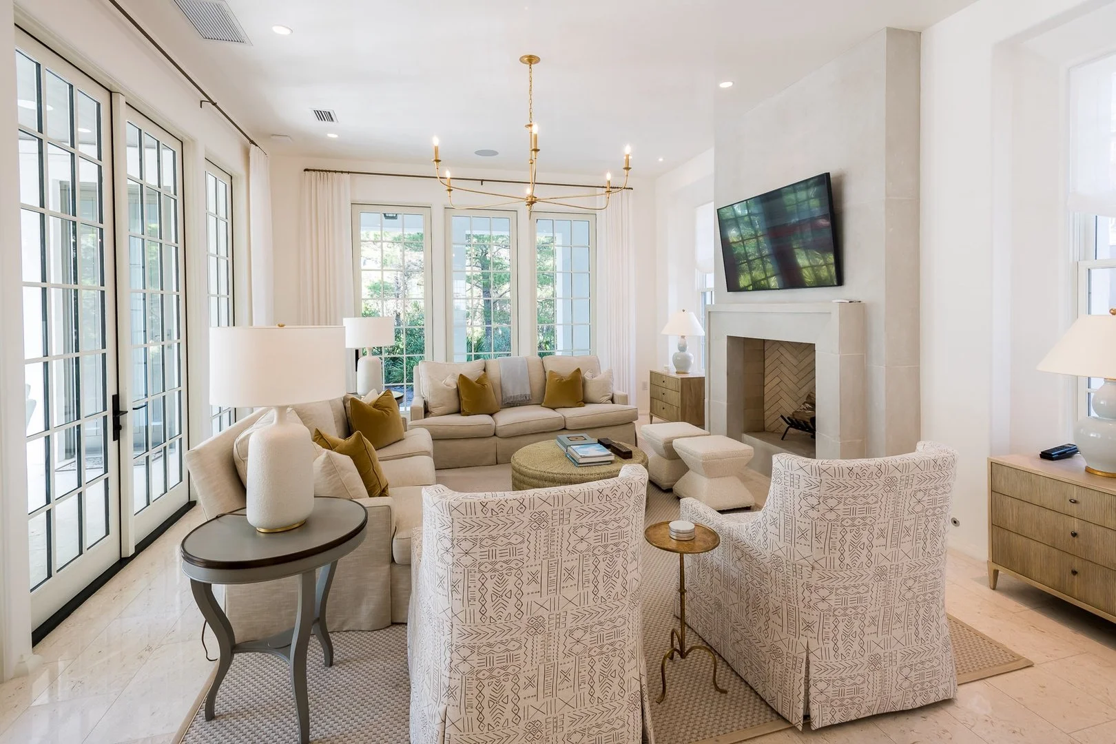 Bright living room with cream-colored sofas, patterned armchairs, a fireplace, large windows providing natural light, and a mounted flat-screen TV.