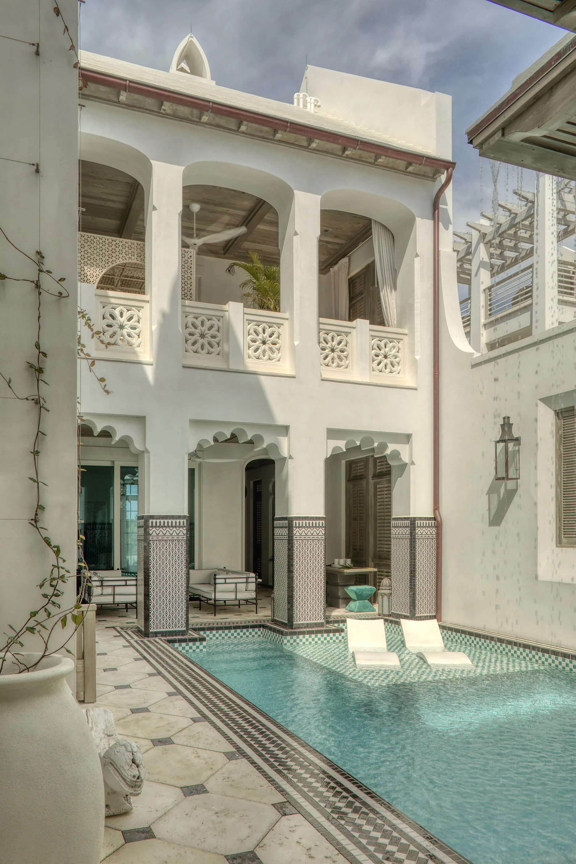 A courtyard with a small swimming pool in front of a white, two-story building with arched windows, decorative balconies, and Mediterranean architecture details. Lounge chairs are in the pool, and there is a seating area with chairs and a small table.
