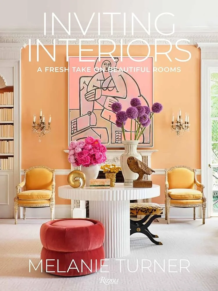 A stylish interior living room with peach walls, two yellow armchairs, a white circular table decorated with pink and purple flowers, a large abstract line art painting, and accents of pink, purple, and gold.