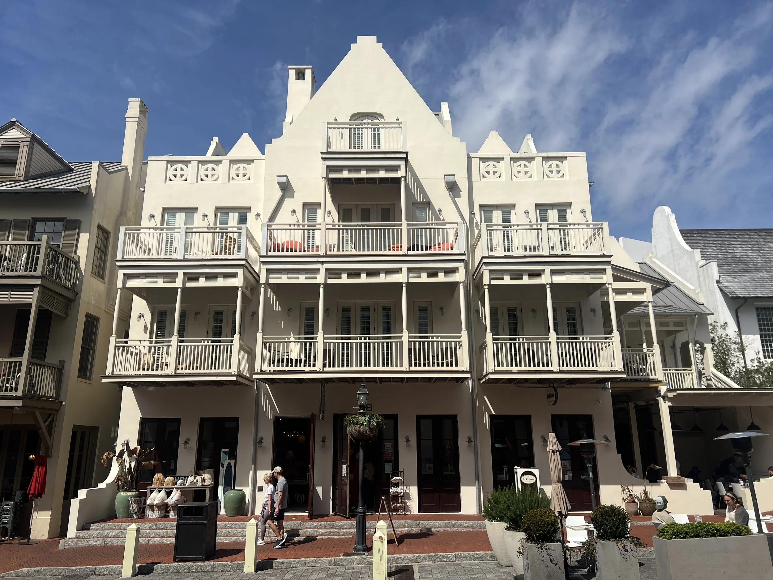 Multi-story white building with balconies and outdoor seating, people walking in front, clear sky.