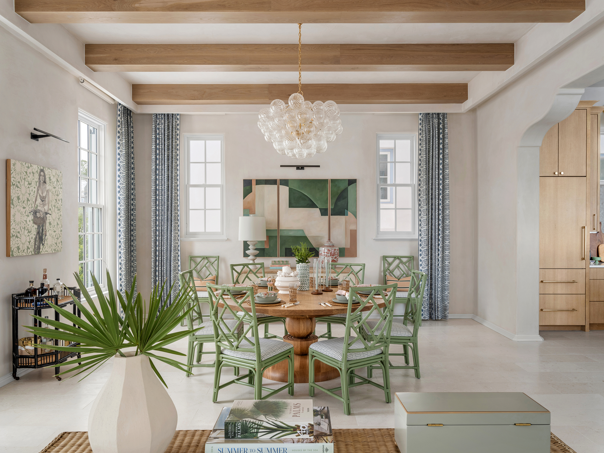 A bright dining room with a round wooden table surrounded by green wicker chairs. The room has a white ceiling with wooden beams, and three windows with patterned blue and white curtains. A chandelier made of glass globes hangs above the table. The t