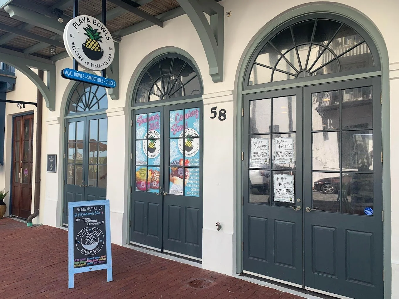Storefront of Playa Bowls with a pineapple logo sign, large green arched windows, and a blue sandwich board sign outside on a red brick sidewalk.
