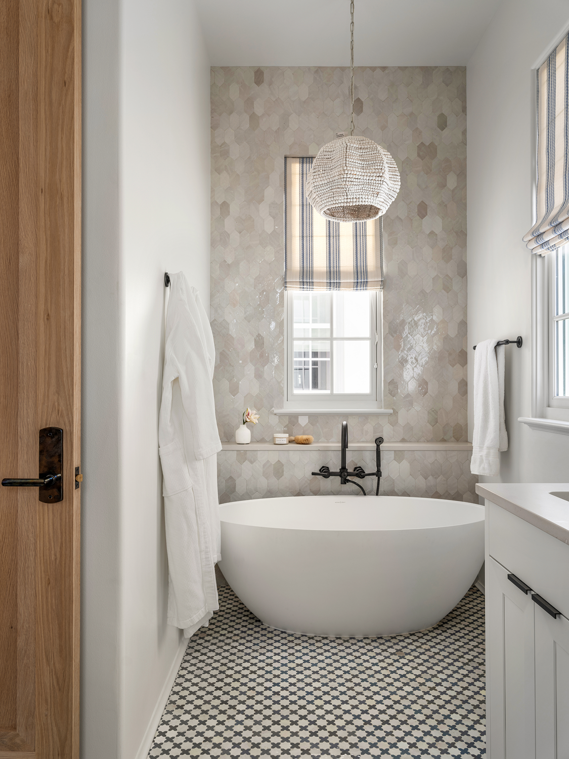 Bathroom with a freestanding tub, patterned floor, window with striped curtain, and textured tile wall.