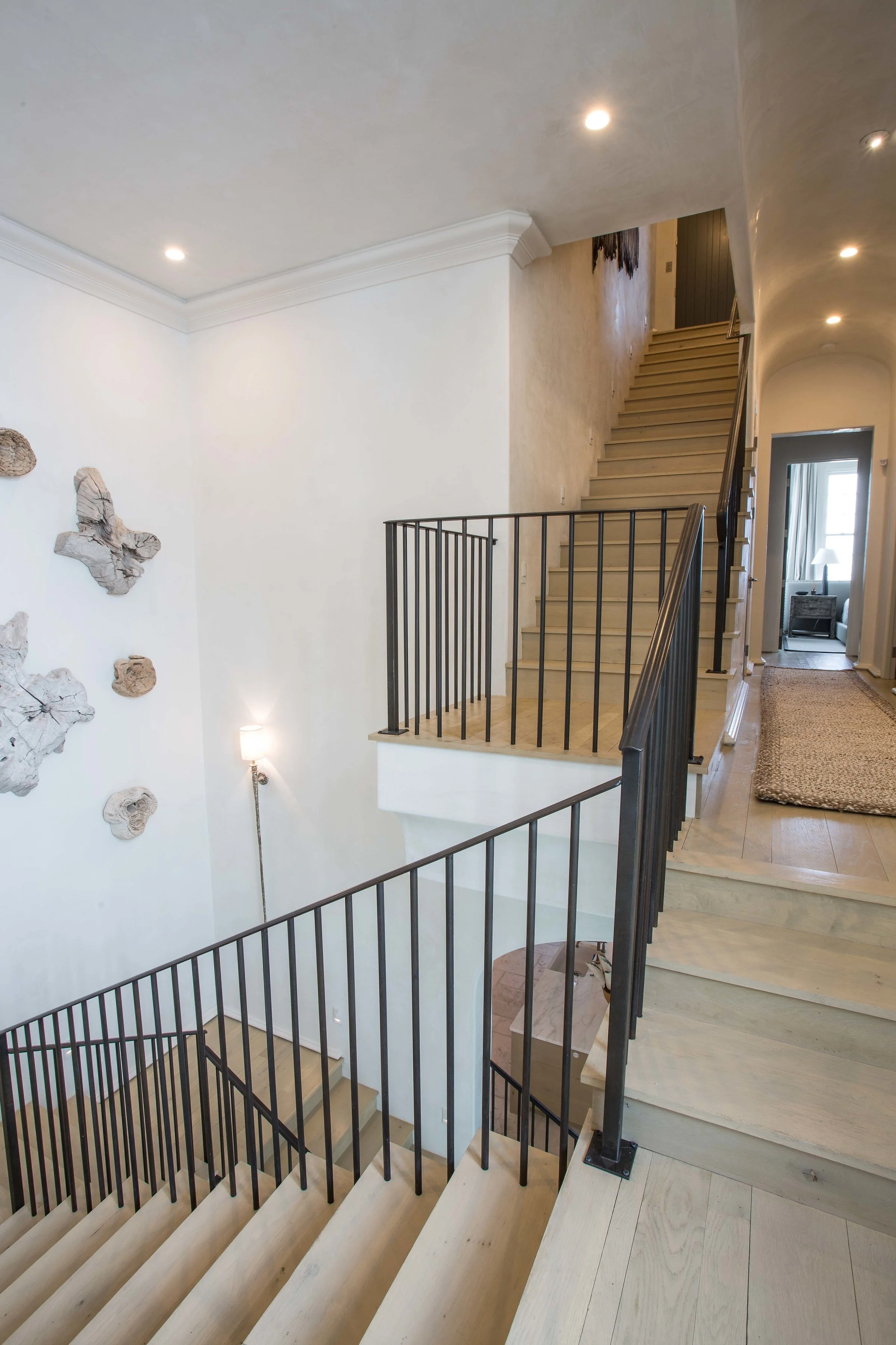 A staircase inside a house, with black metal railing, light-colored wooden steps, white walls, and some wall art, leading to an upper floor and hallway with natural light.