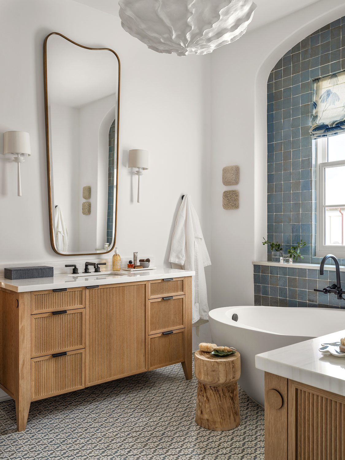 Modern bathroom with white walls, a large mirror, wood vanity with white countertop, black faucet, bathtub near blue tiled window, and decorative elements like a cloud-shaped ceiling fixture and textured art pieces on the wall.