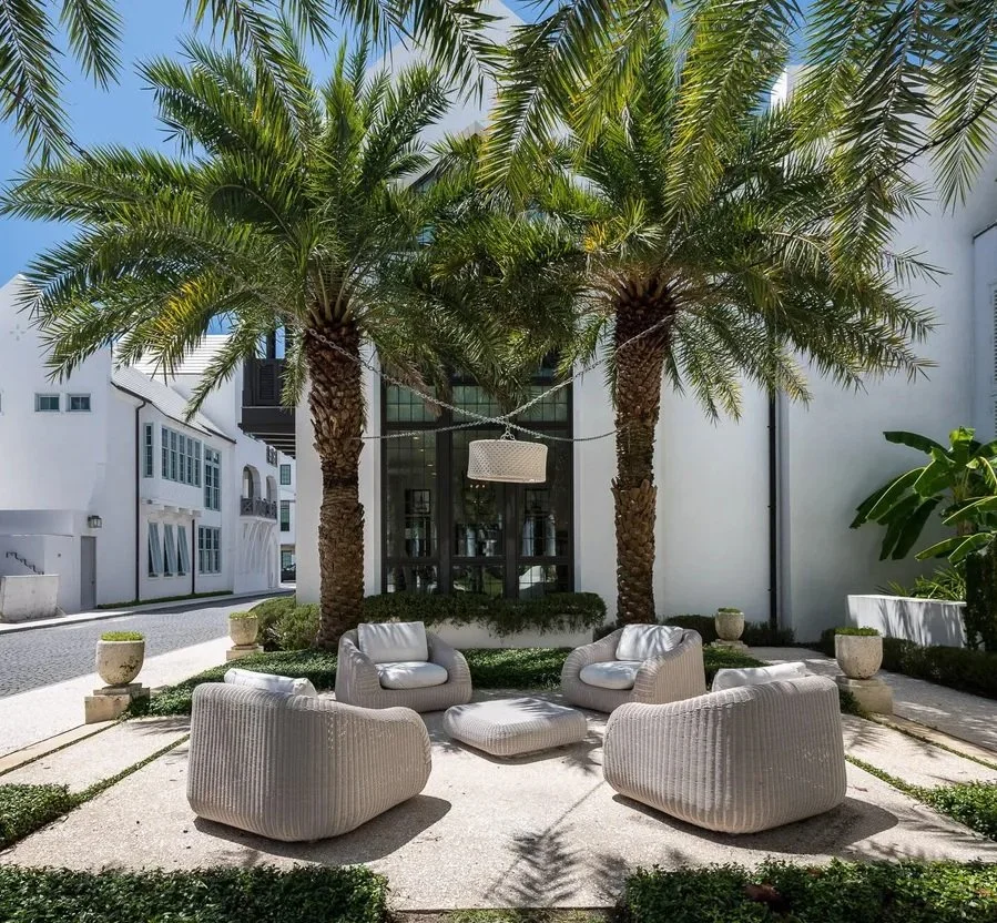 Outdoor seating area with four modern beige armchairs and an ottoman under two large palm trees, with white planters and greenery surrounding the space.