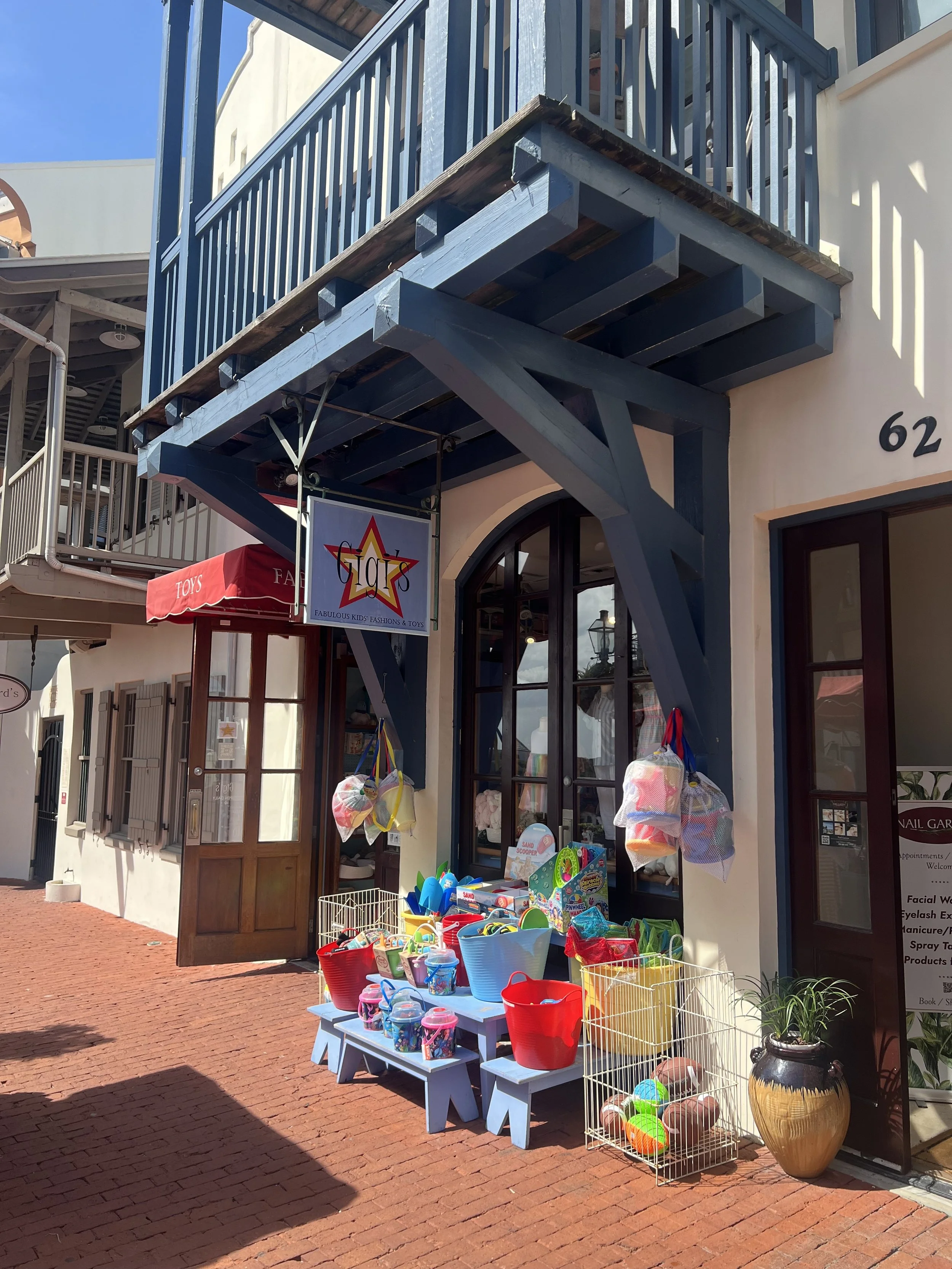 Children's toy shop with outdoor display of buckets filled with sand toys, colorful balls, and other toys on blue benches and a white wire rack on a brick sidewalk.