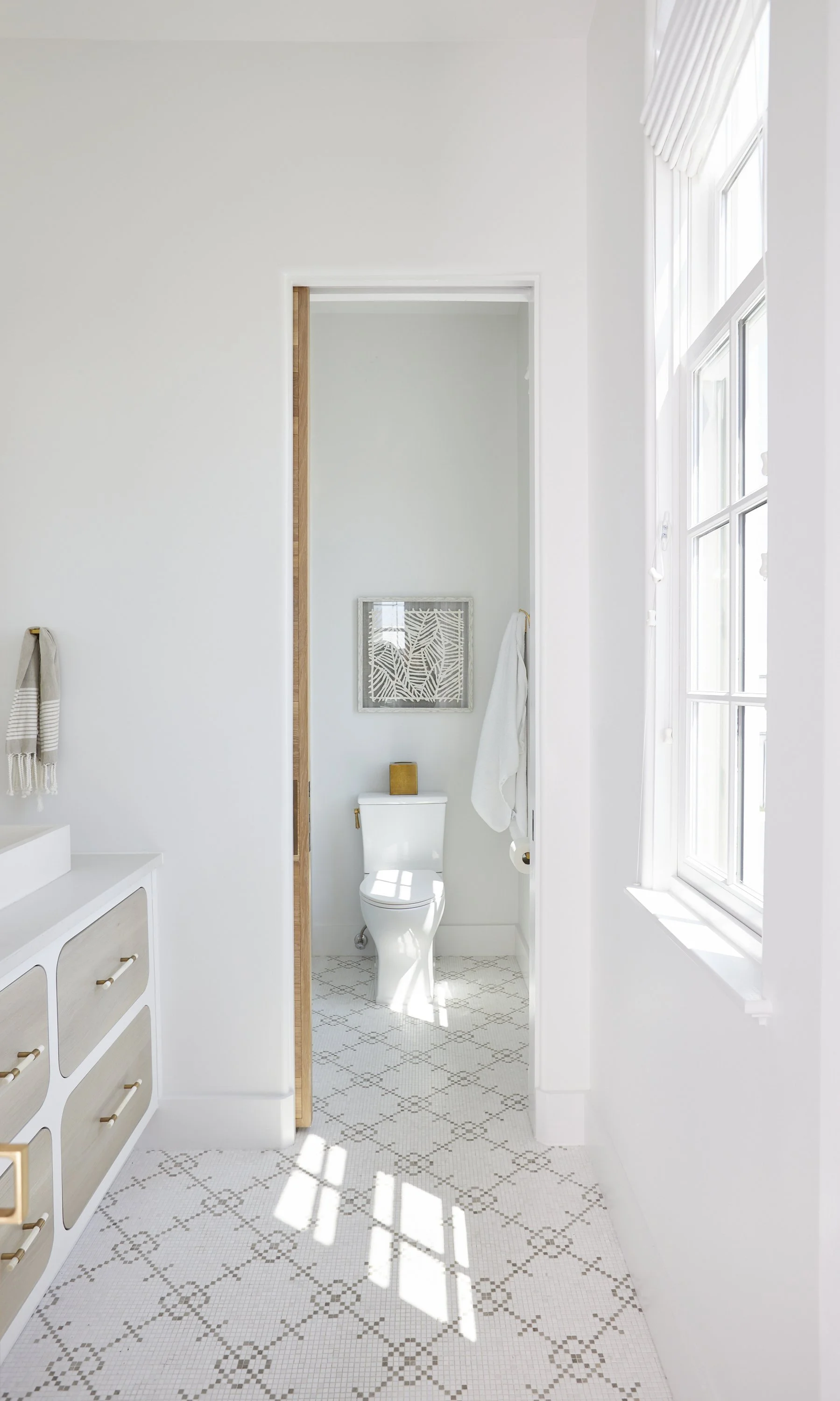 Bright white bathroom with a toilet, window with sunlight, and light-colored patterned tile floor.