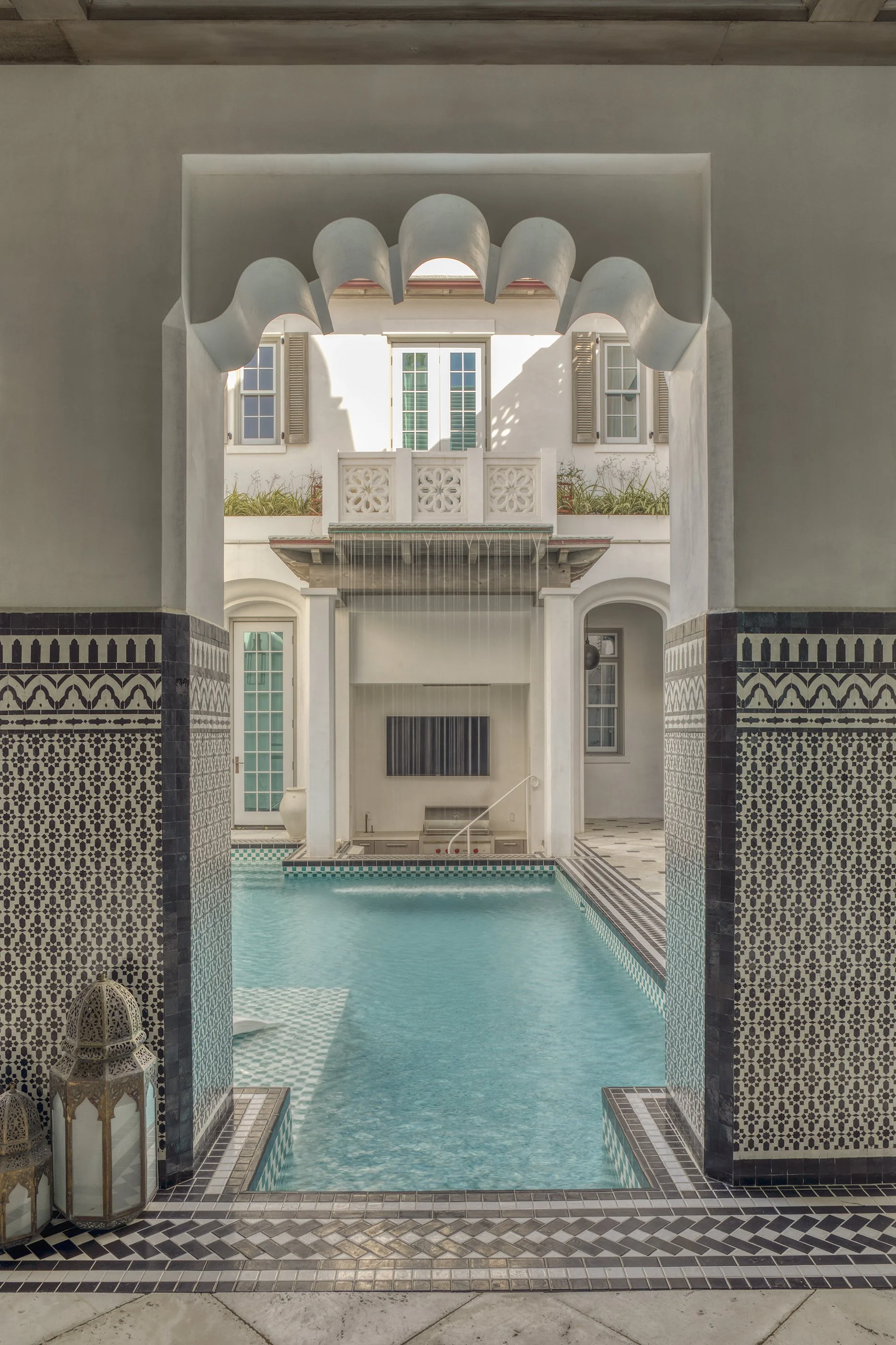 Indoor swimming pool with Moroccan-style tile work, viewed through an archway with scalloped edges, overlooking a sunlit courtyard with a second-story balcony and windows.