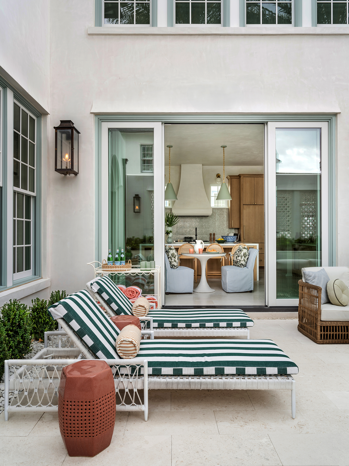Outdoor patio with three striped lounge chairs, a small red side table, and a wicker sofa, leading into a kitchen and dining area through large glass sliding doors.