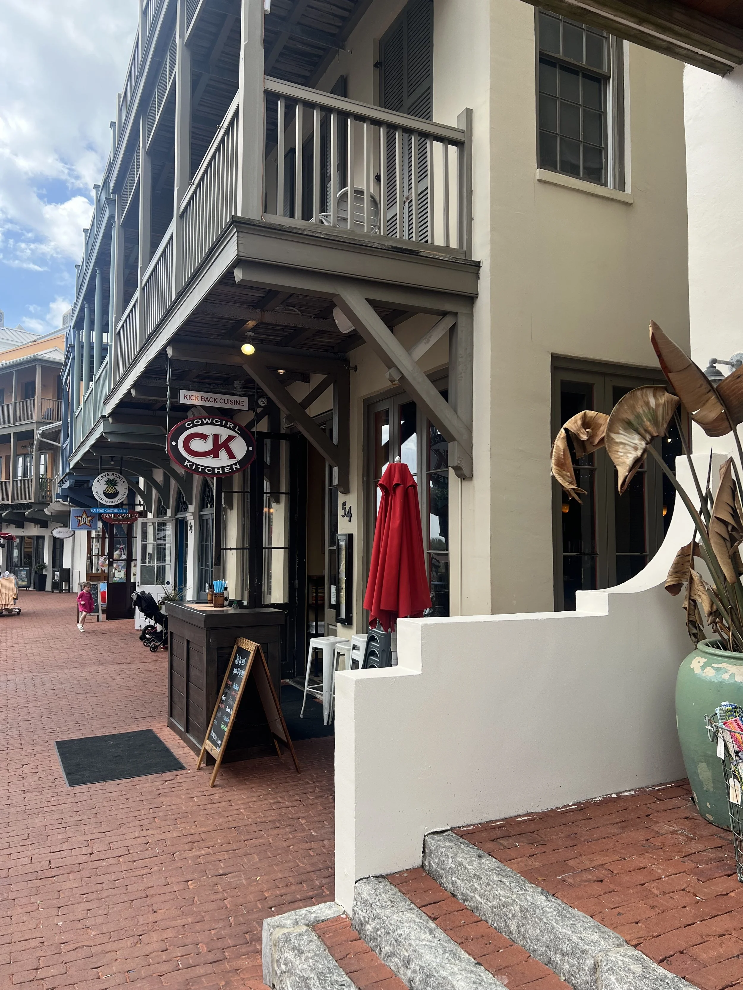 Exterior of a restaurant called Cowgirl Kitchen with a sign reading 'Kick Back Cuisine.' There is a chalkboard menu outside, a red umbrella, and an outdoor seating area. Brick sidewalk and a person in the background.