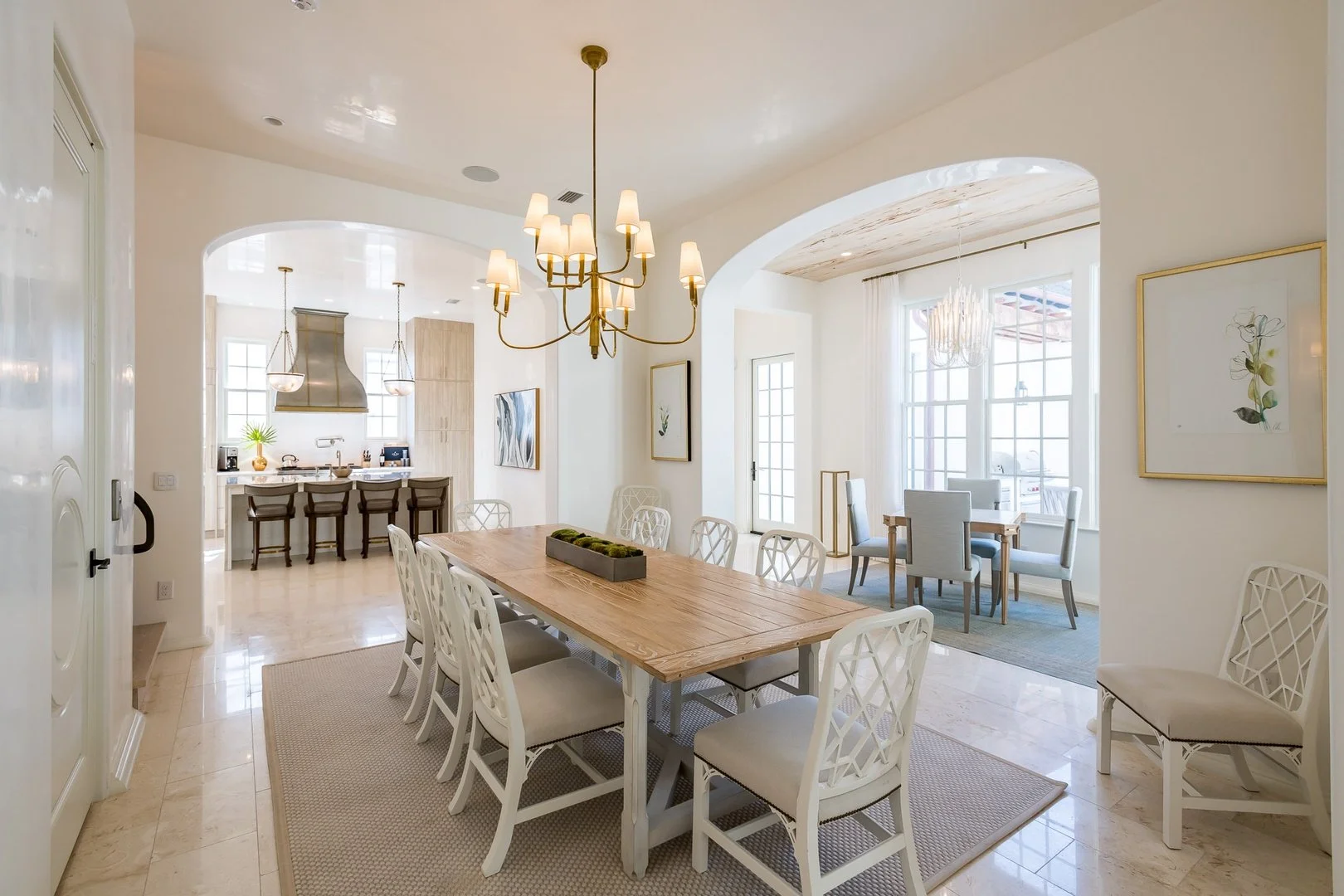 Interior of a bright, airy dining room with a long wooden table surrounded by white chairs, a chandelier overhead, and a cozy sitting nook with a table and four chairs near large windows.