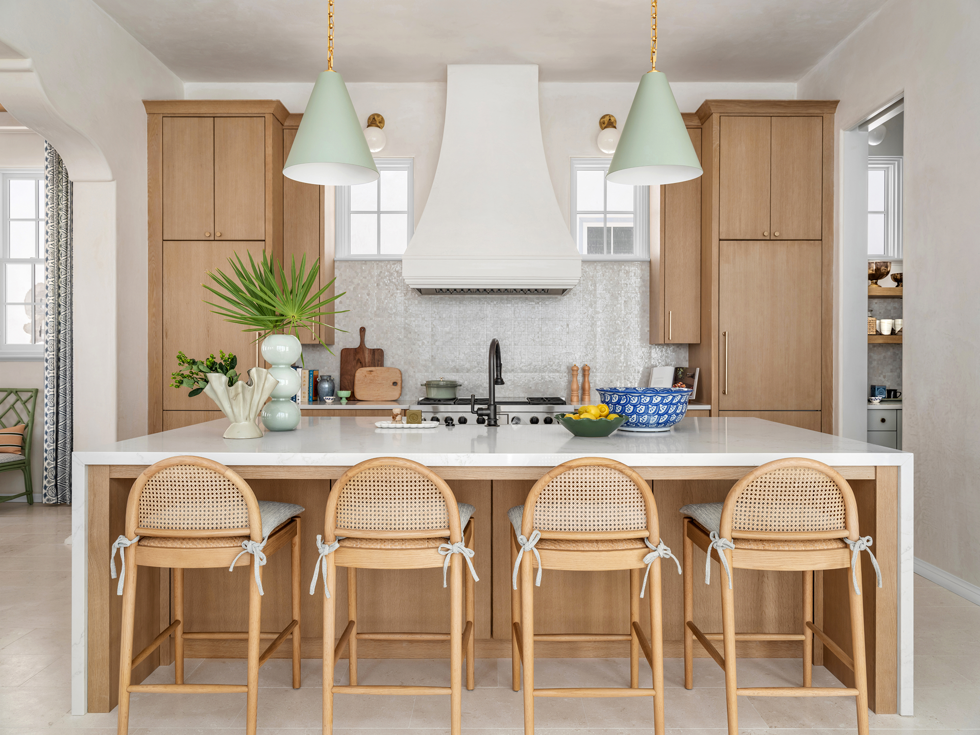 Modern kitchen with light wood cabinets, white walls, and a white marble island with four beige rattan bar stools tied with light blue bows.