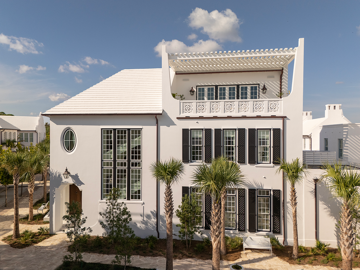 White coastal-style house with black shutters, large windows, and a second-floor balcony with decorative railing, surrounded by palm trees under a blue sky.