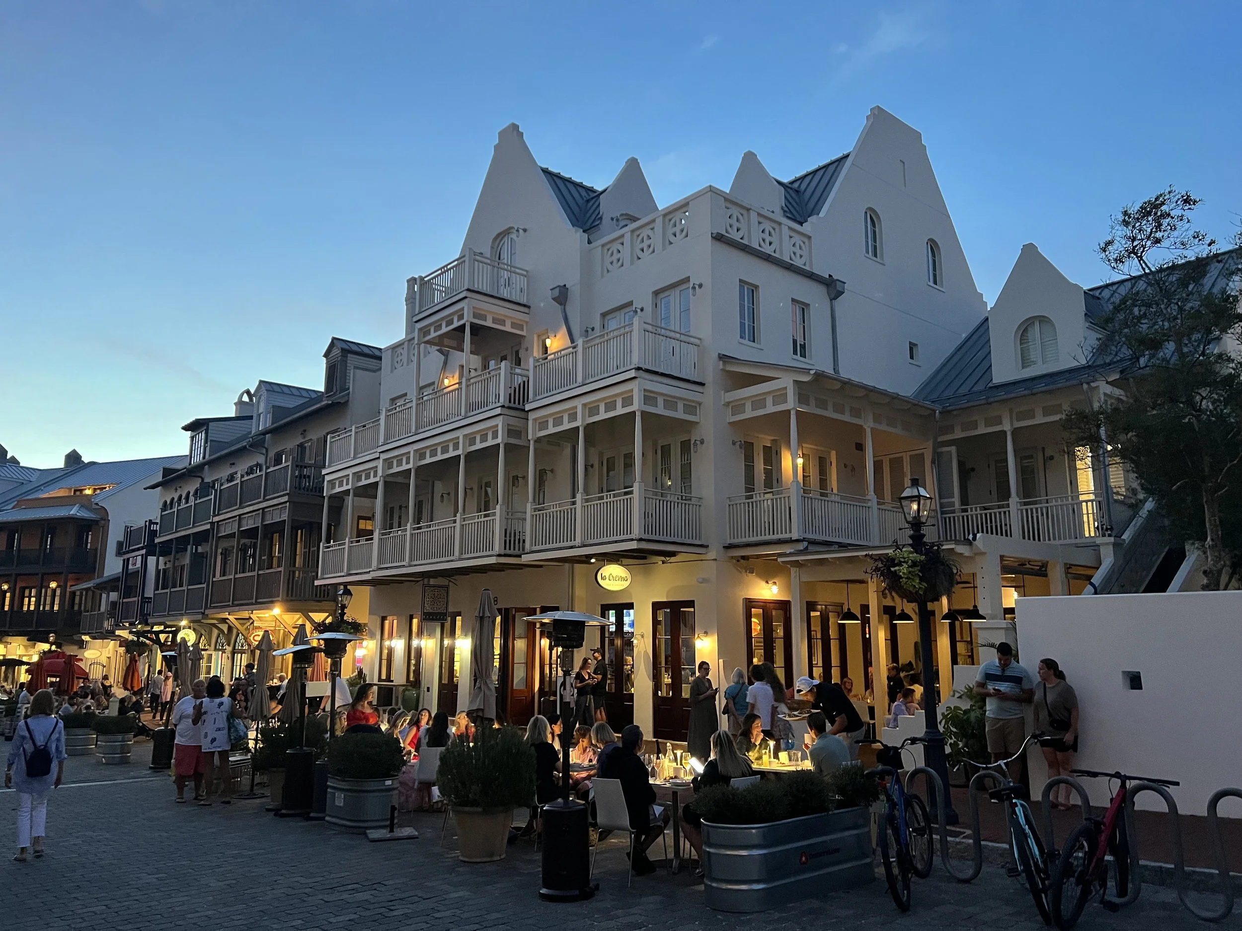 A lively street scene at dusk with people dining outside a multi-story building resembling a small castle, with balconies, with some lit windows, and bicycles parked in the foreground.