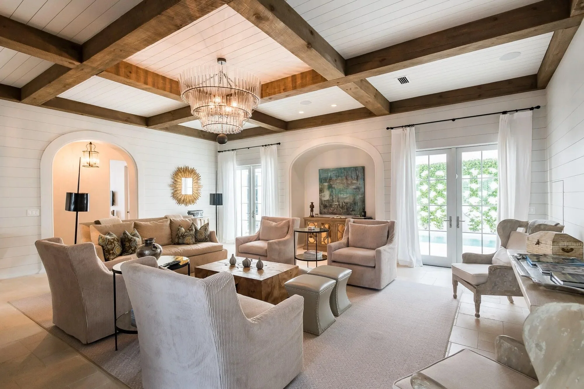 Living room with white walls, beige furniture, wooden ceiling beams, chandelier, French doors with white curtains, and decorative decor.