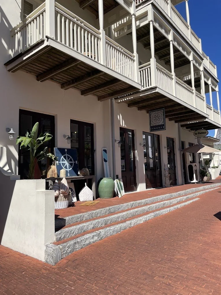 Storefront with outdoor decor and furniture, including a large green vase, baskets, and nautical-themed items, under a balcony with white wooden railings on a sunny day.