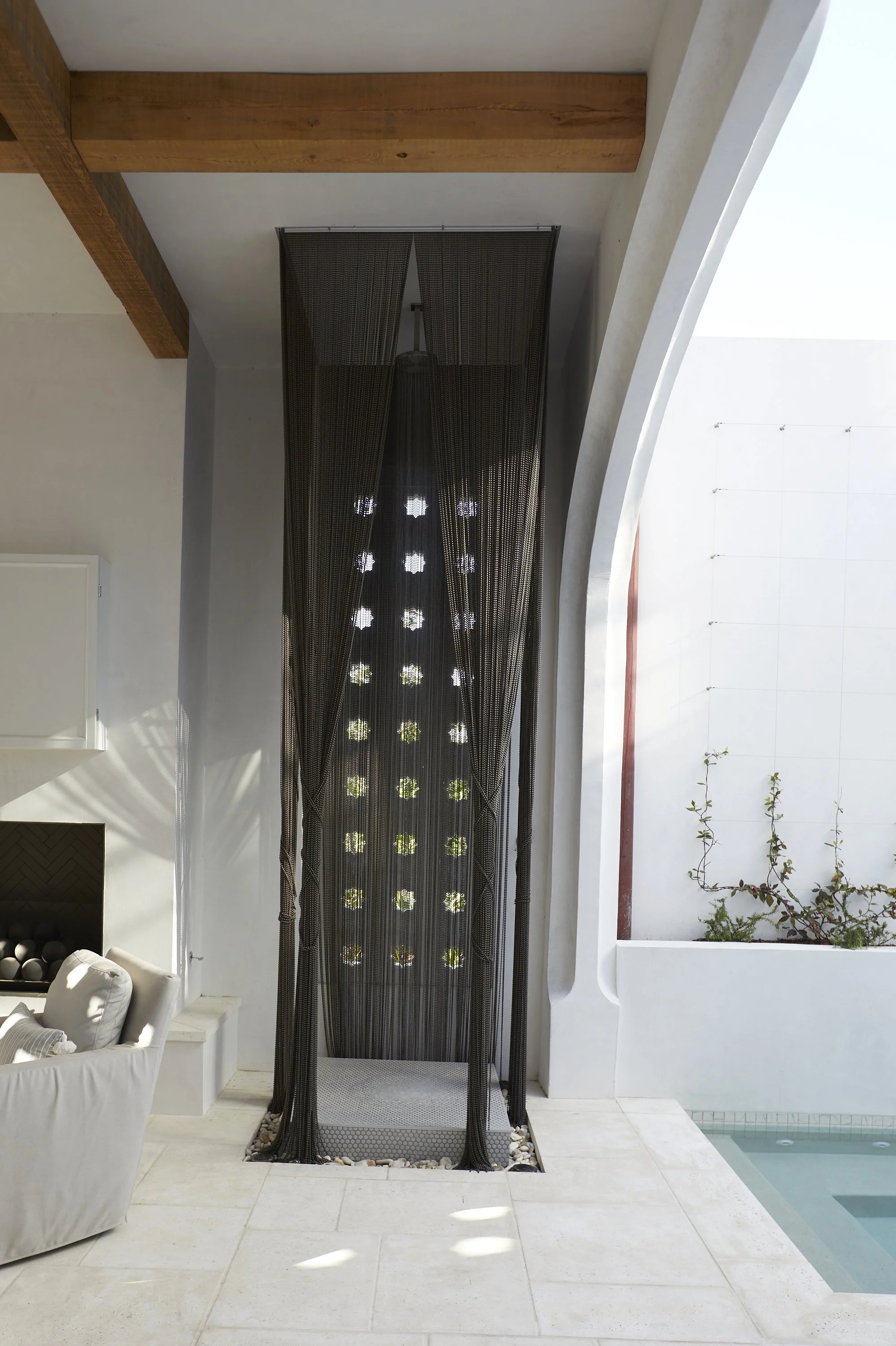 An indoor shower with black beads hanging as a curtain, situated next to a poolside area with white walls and some greenery.