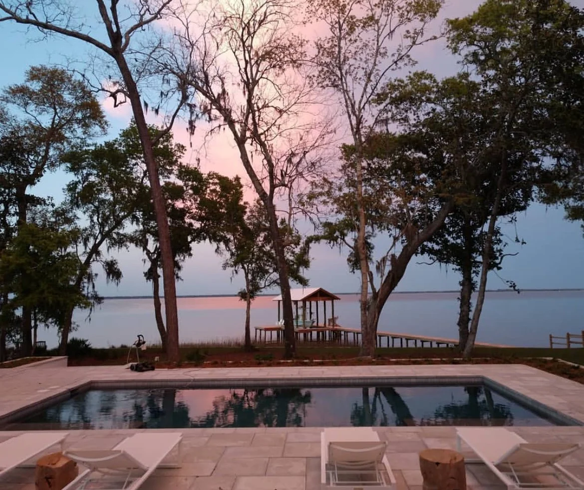 A tranquil lakeside scene at sunset with a pool in the foreground, surrounded by lounge chairs, trees, and a dock leading to a gazebo over the water.