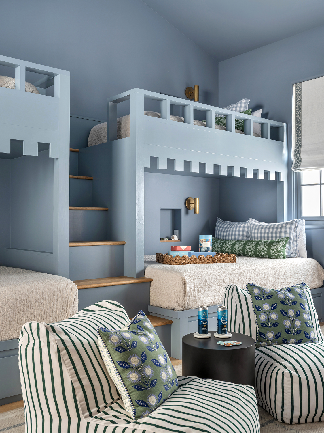 A cozy indoor space with a built-in bunk bed, featuring blue walls, striped and floral pillows, a window with a roller shade, and a small table with drinks.