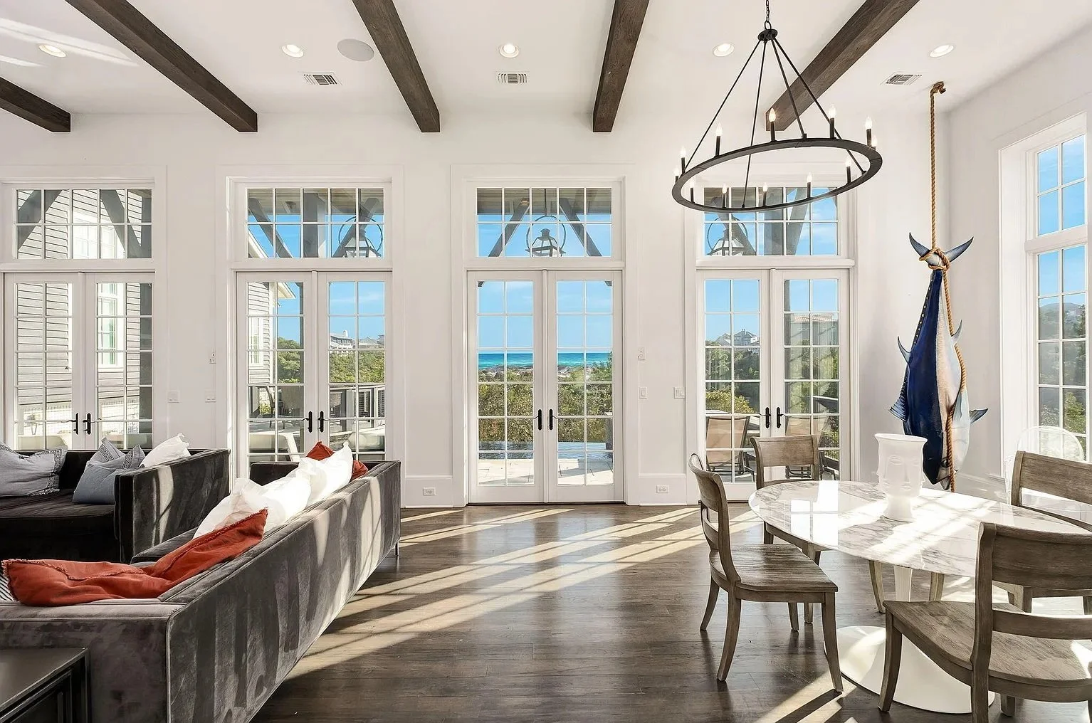 Living and dining room with large windows, a view of the ocean, wooden beams on the ceiling, a chandelier, a sectional sofa, pillows, a round marble dining table, chairs, and an American flag and fish mounted on the wall.