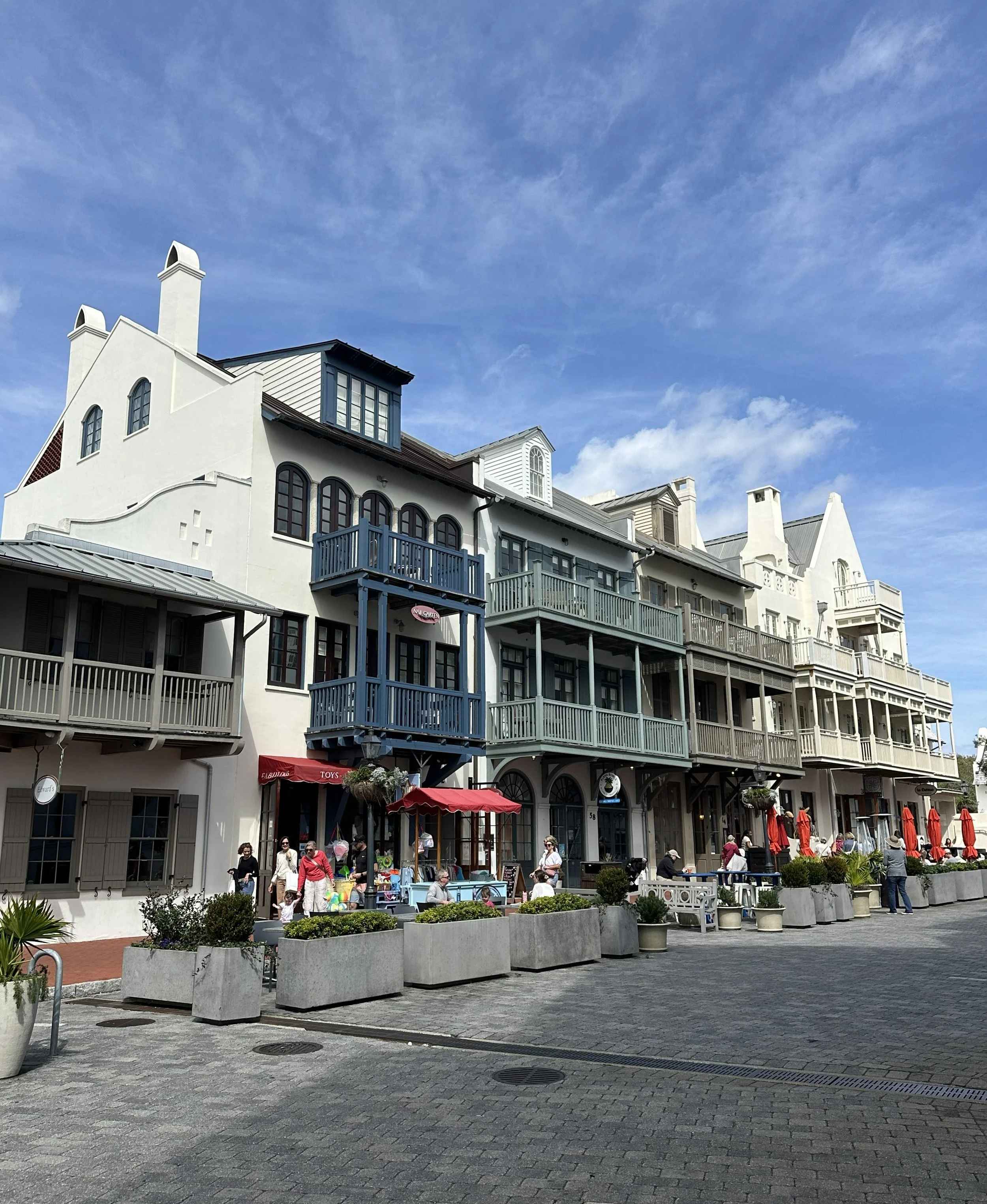 People walking and sitting outside a colorful, multi-story building with shops and cafes under a partly cloudy sky.