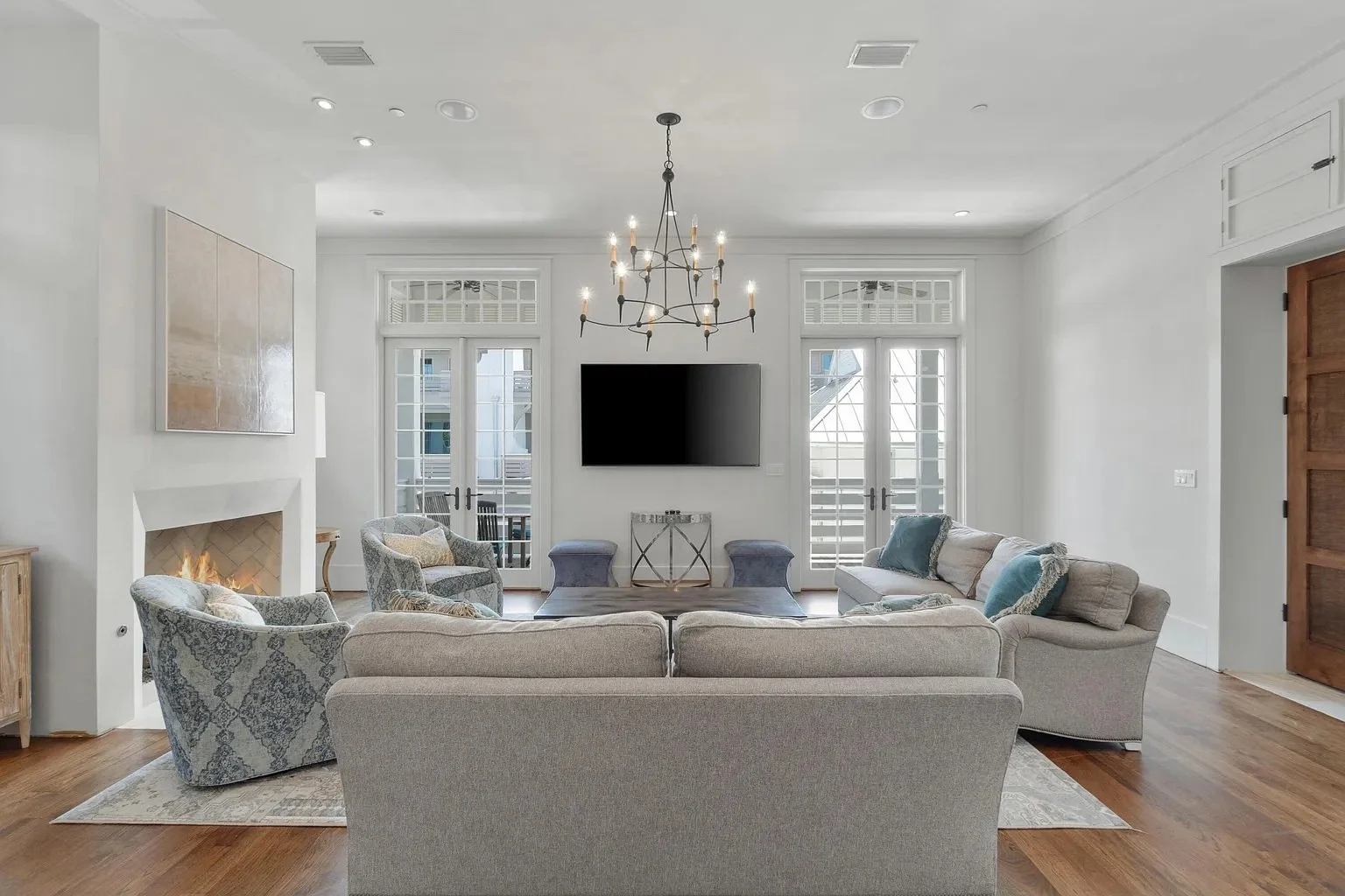 Bright living room with beige sofas and patterned armchairs around a coffee table, fireplace on the left, large flat-screen TV on the wall, and French doors leading to the balcony, decorated in neutral tones with a chandelier hanging from the ceiling