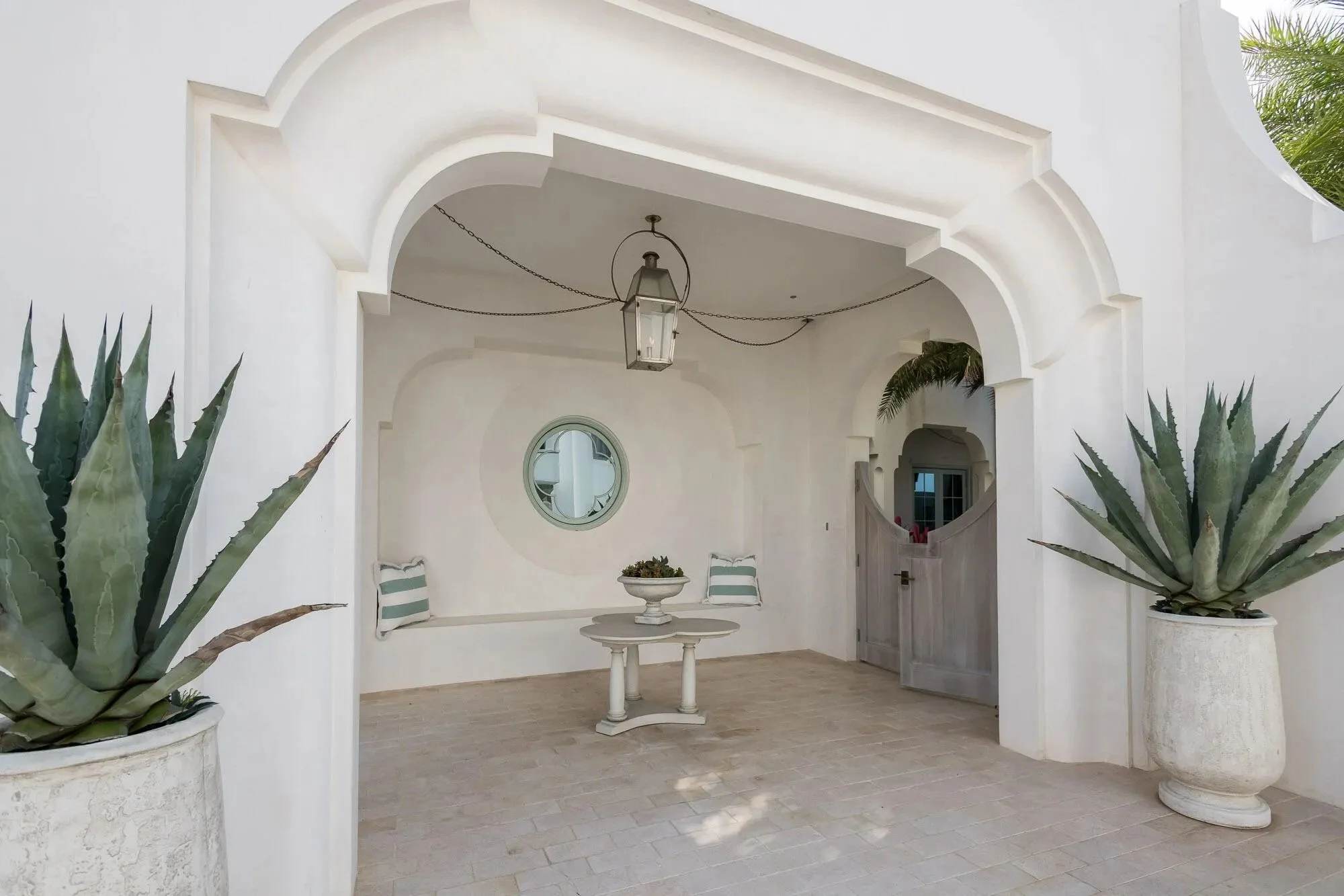White covered patio with large potted agave plants, a round table with a planter, a bench with cushions, a circular window, and a hanging lantern