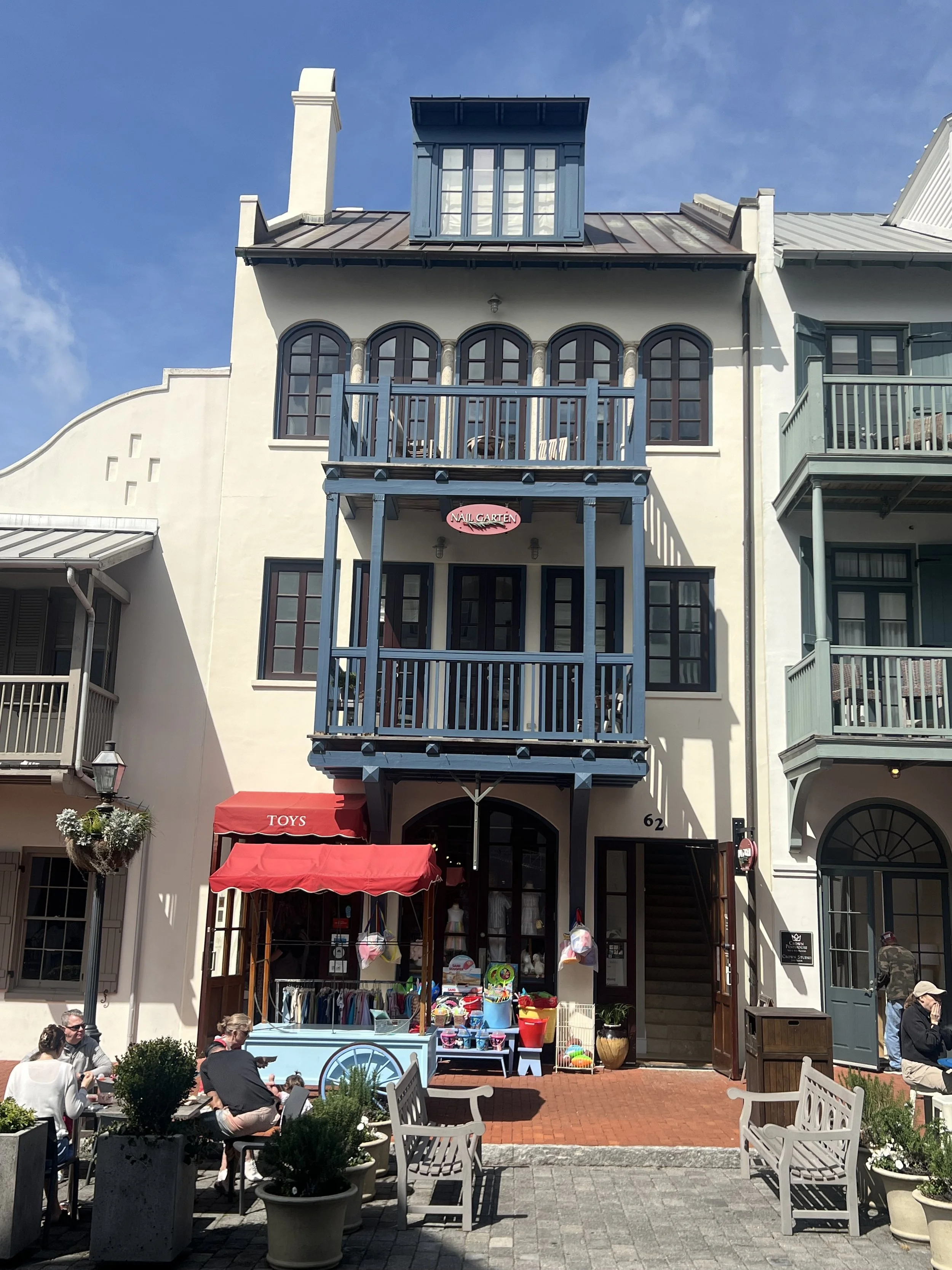 A three-story building with a blue balcony and a rooftop structure. The ground floor has an outdoor toy shop with red awning and people sitting at tables outside. There are potted plants and a brick sidewalk in front.