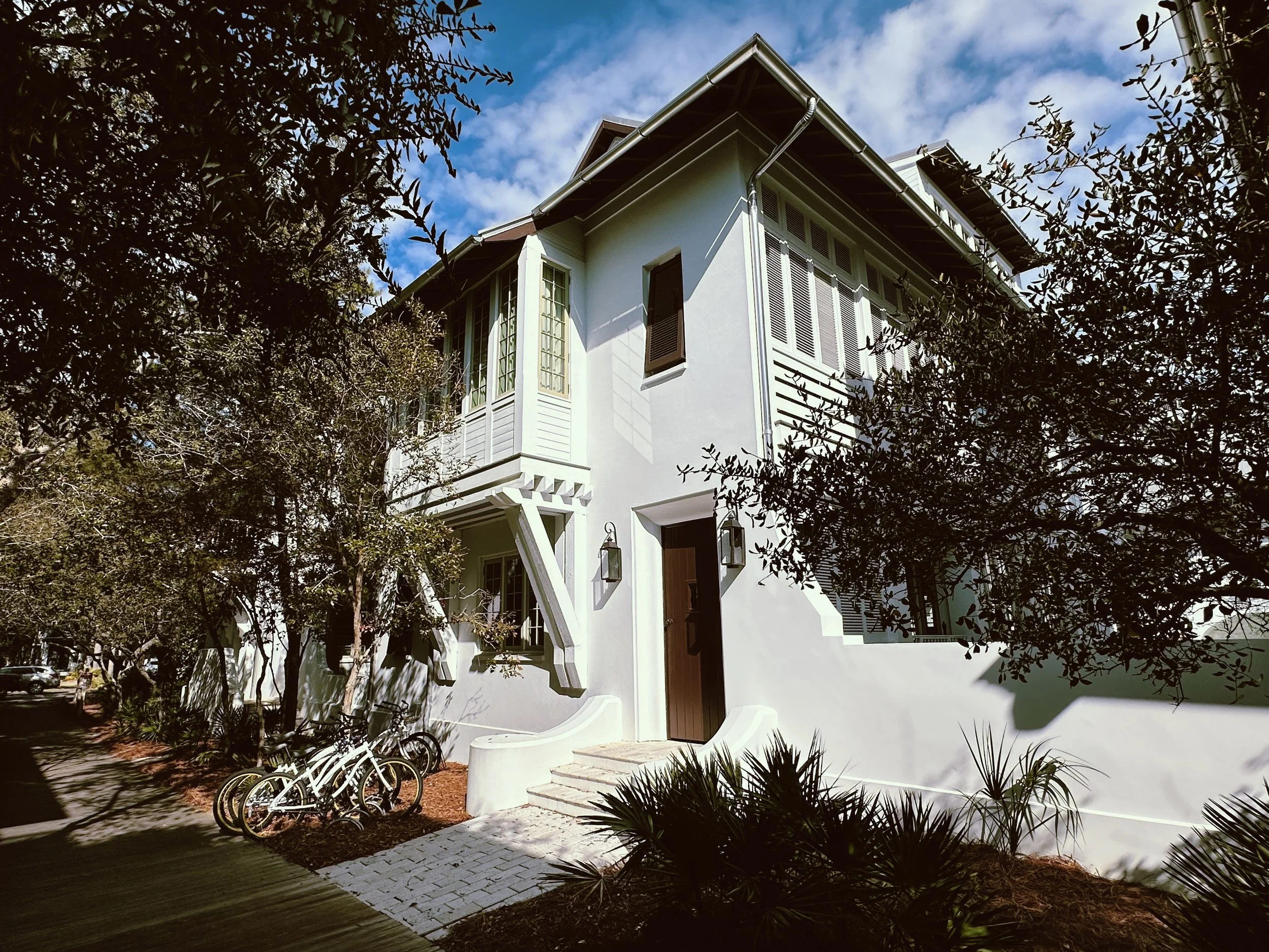A two-story white house with dark brown front door, surrounded by trees and plants, with bikes parked outside and a blue sky overhead.