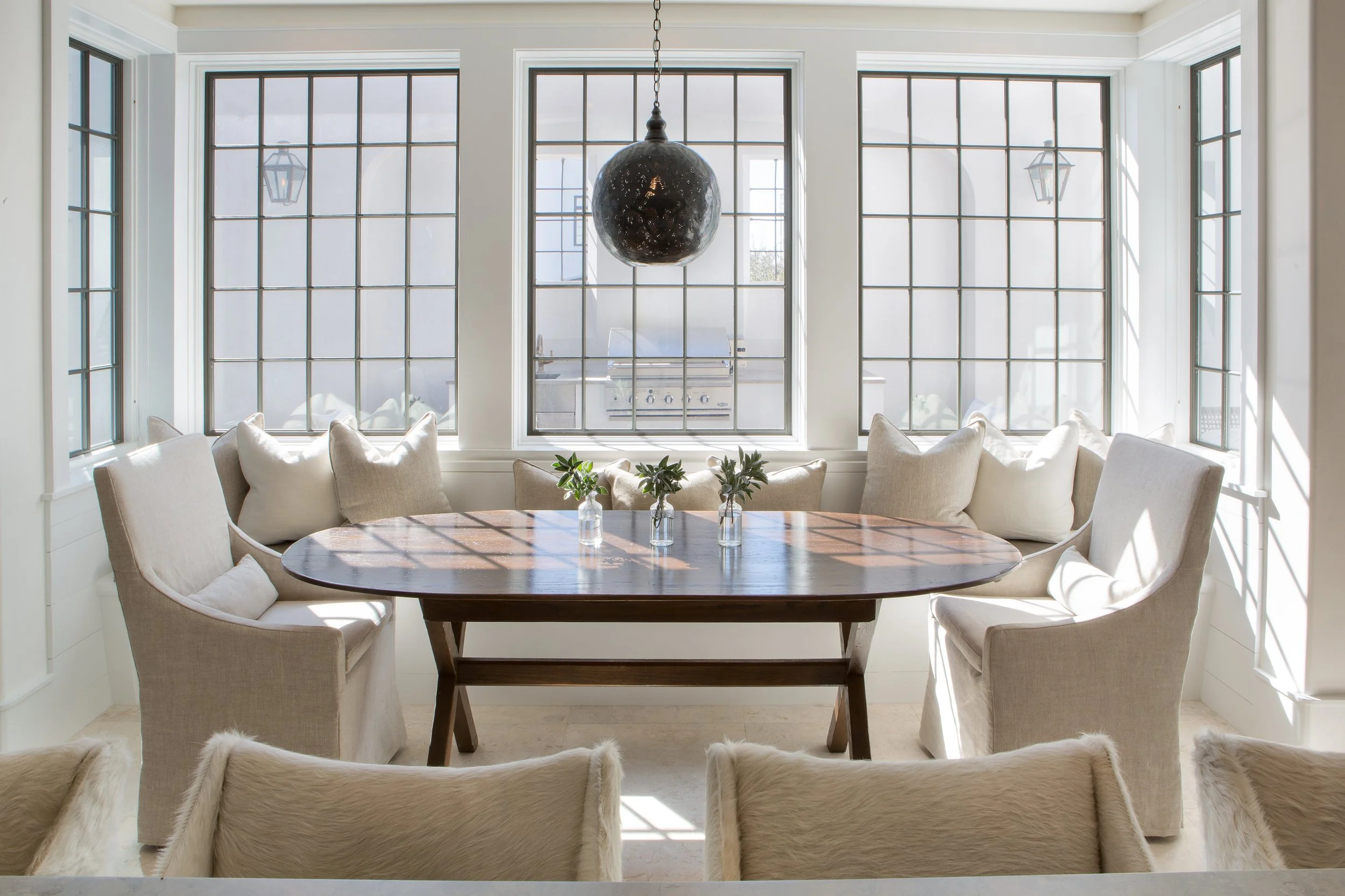 Bright dining room with large windows, a wooden table, beige upholstered chairs, and small potted plants on the table. A dark pendant light hangs above the table, and there is a grill visible outside through the windows.