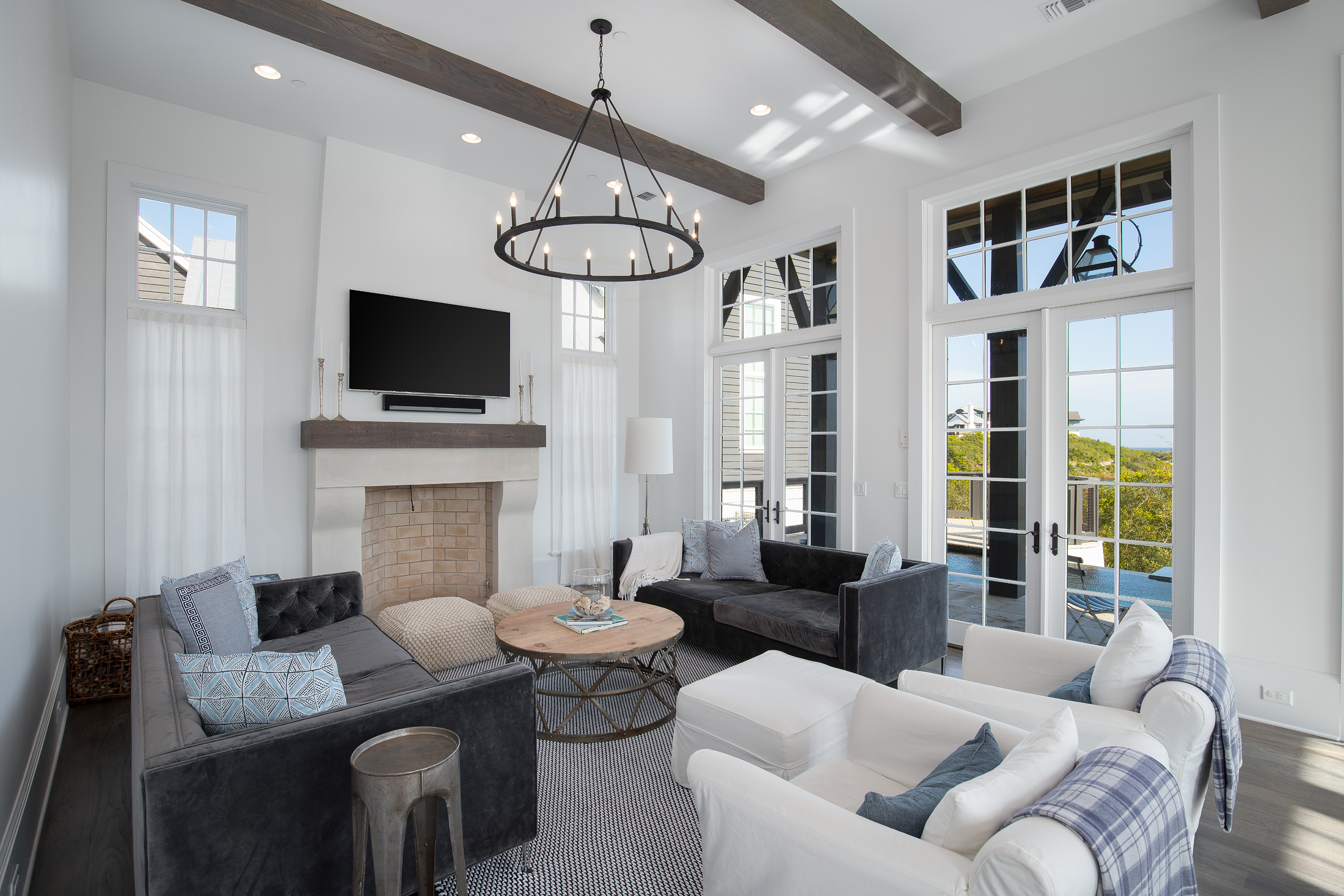 Modern living room with white walls, large windows and French doors offering a view outside. There are two black sofas, a white armchair, a round wooden coffee table, and a fireplace with a stone interior. A large circular black chandelier hangs from