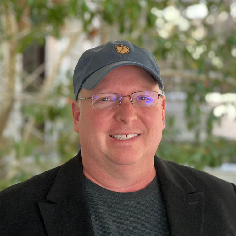 A smiling man wearing glasses, a dark jacket, and a baseball cap outdoors with blurred green foliage in the background.
