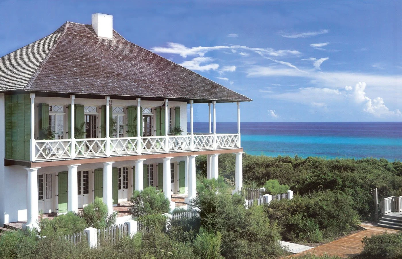 A large, two-story beach house with a wrap-around porch, white columns, and green shutters, overlooking the ocean with a clear blue sky and some clouds.