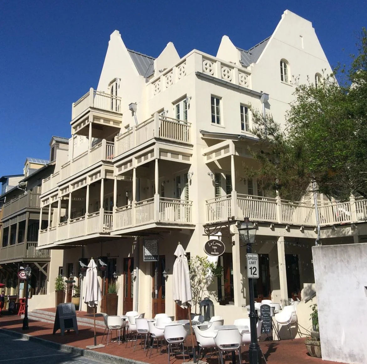 A white, multi-story building with balconies and outdoor seating, houses a restaurant called La Crema in a lively street with outdoor tables and umbrellas.