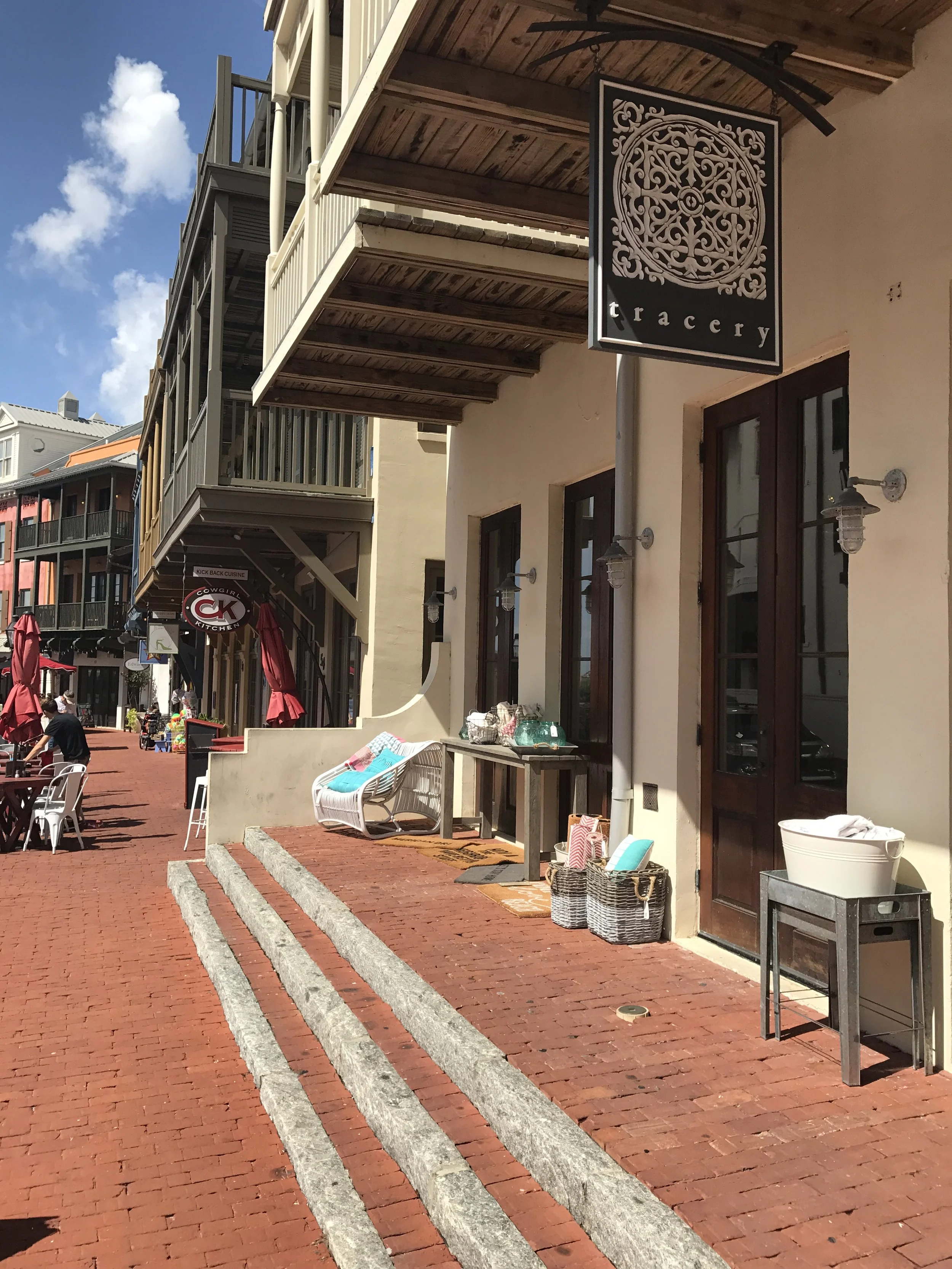 Street view of a sidewalk with outdoor seating for a cafe or shop, featuring a sign that reads 'tracery,' and various baskets and chairs outside the storefronts. Buildings with balconies line the street under a blue sky with some clouds.