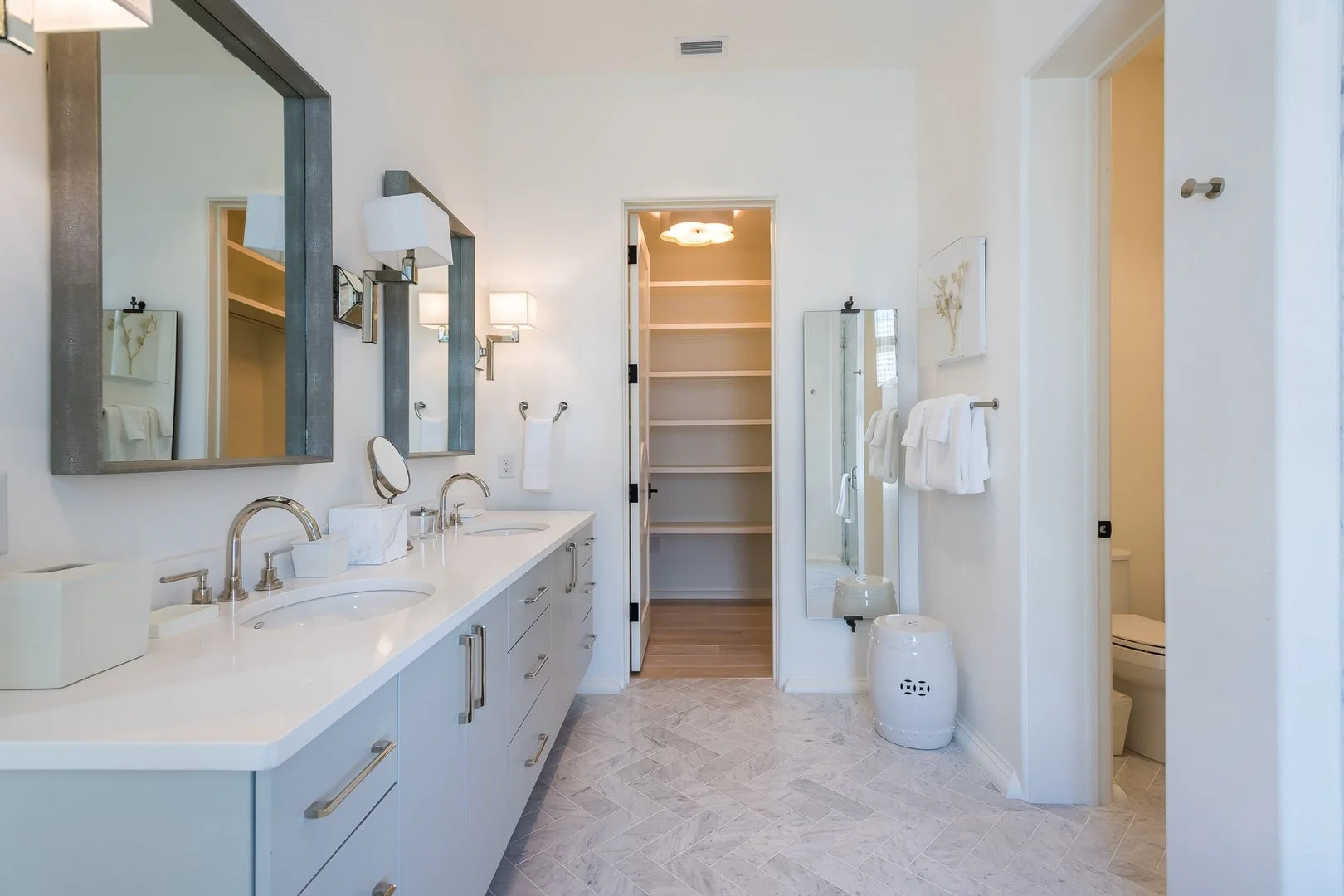 A modern, bright bathroom with a white double vanity, large mirrors, and wall-mounted light fixtures. There are towels hanging on the wall, a full-length mirror, and a walk-in closet with shelves. A toilet is partially visible in a small room to the 