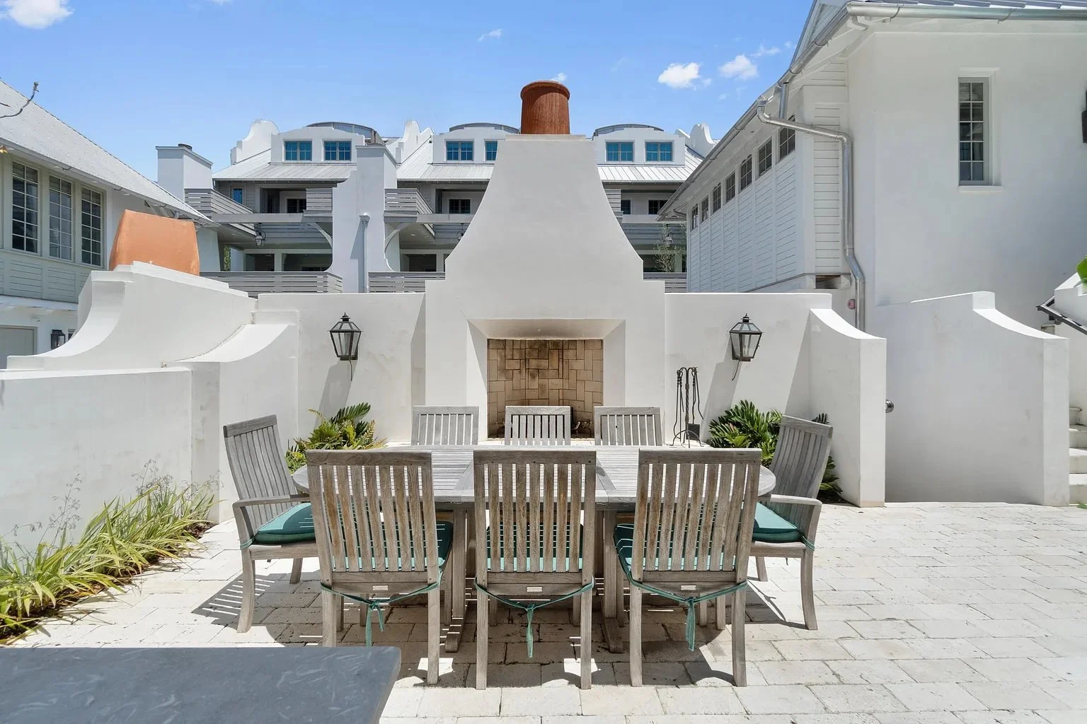 Outdoor patio with dining table and chairs, white walls, fireplace, and lanterns under a blue sky.