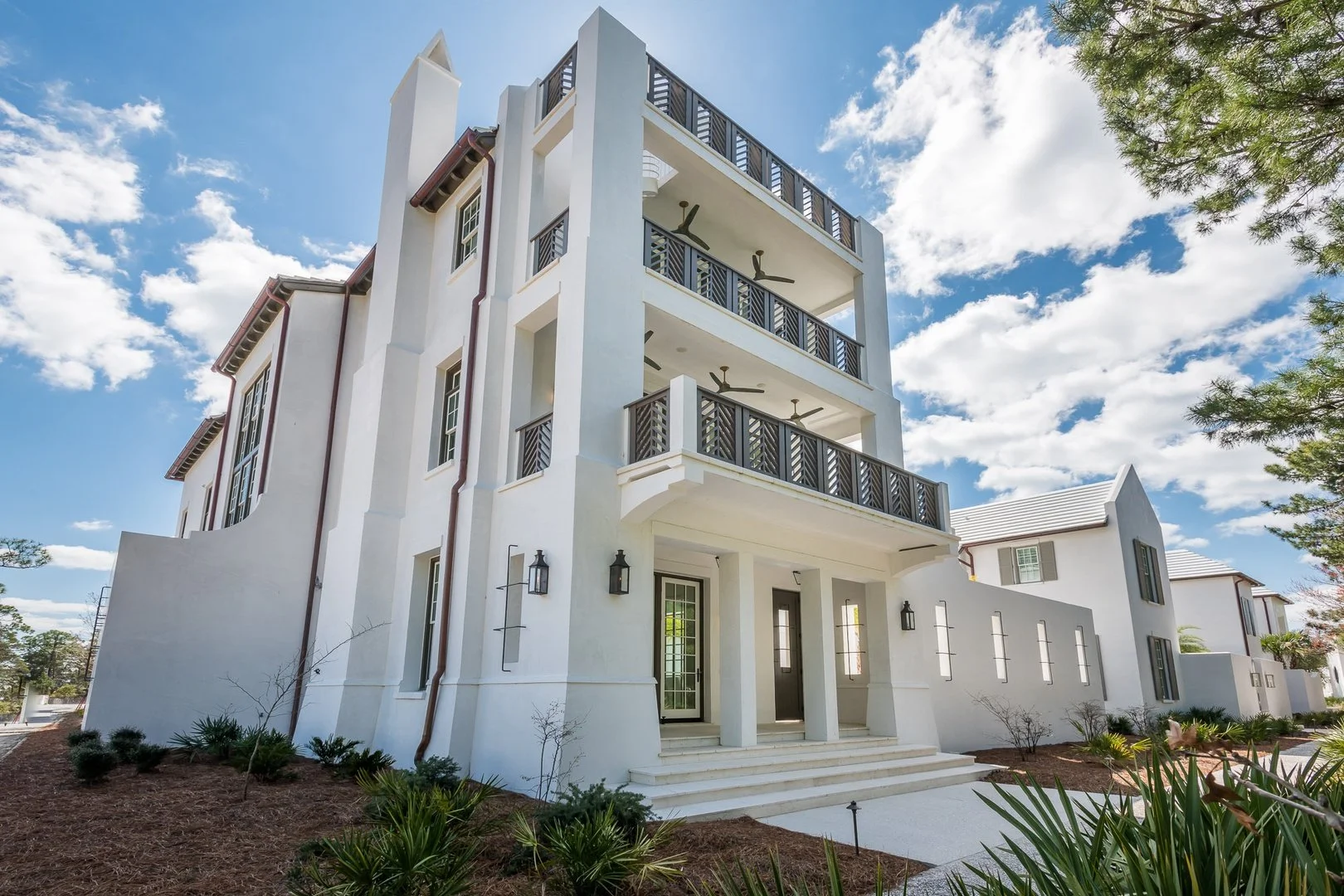 A modern, white multi-story house with balconies, black railings, and ceiling fans, set against a bright blue sky with scattered clouds.
