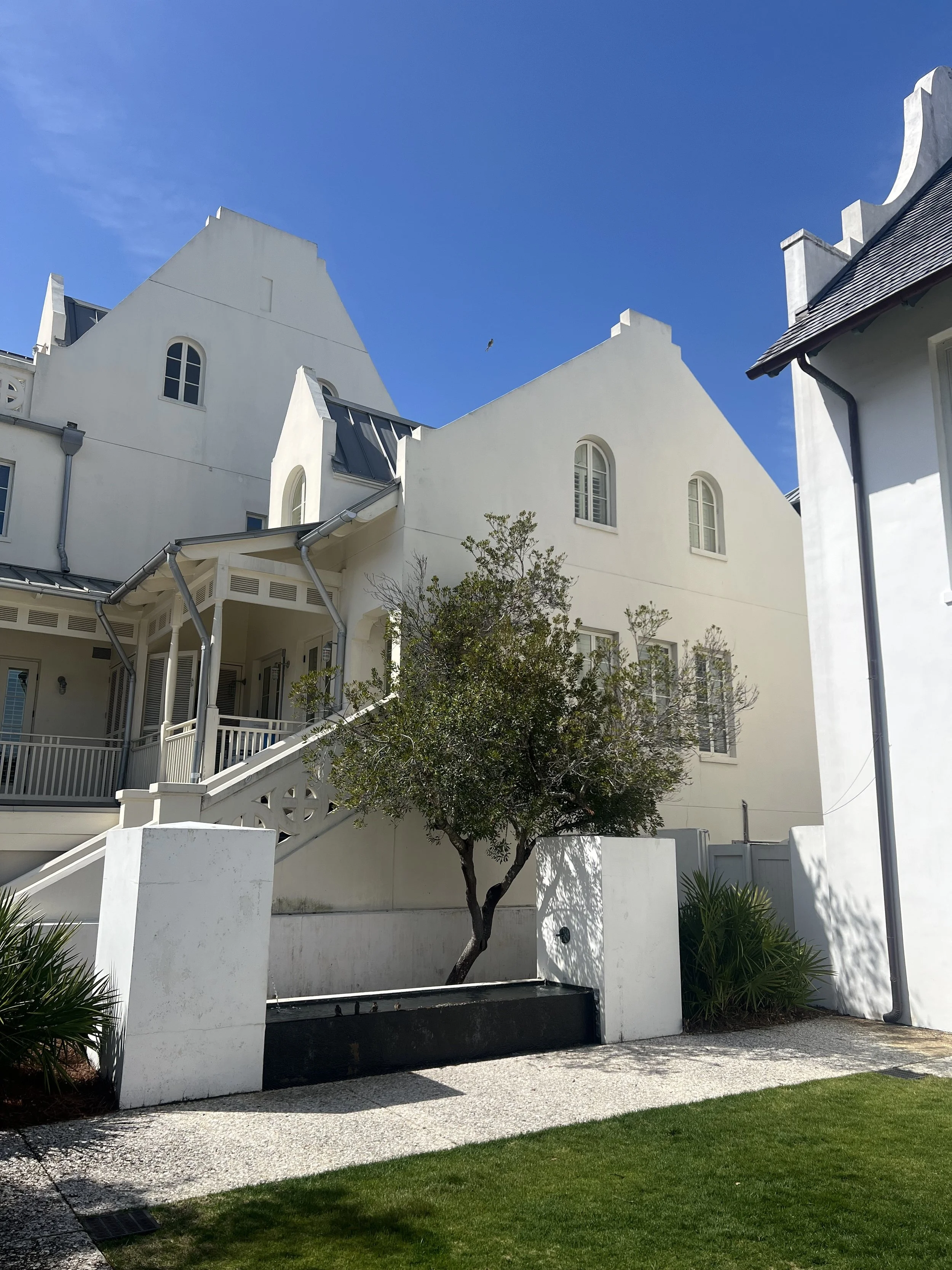 White multi-story house with gabled roofs, a small tree in front, and a clear blue sky.