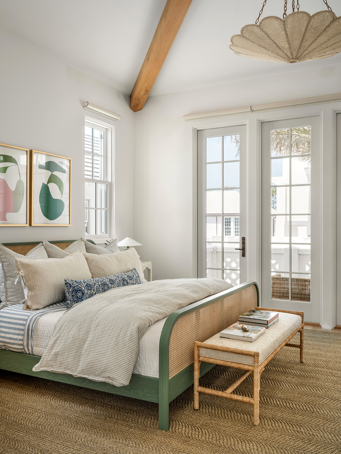 A bright bedroom with a green bed, decorated with multiple pillows, and a rattan bench at the foot of the bed with books on top. Large windows and a glass door lead to a balcony, with artwork on the wall and a ceiling light fixture visible.